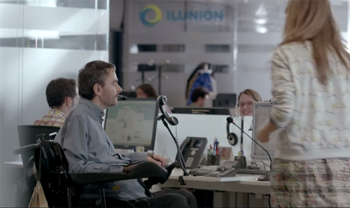 
En una oficina de Ilunion, personas con y sin diversidad funcional trabajan en sus puestos. Un chico con movilidad reducida charla con una compañera sentado en su silla de ruedas, frente a un ordenador y un teléfono adaptados.
