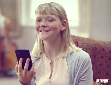 
Una chica joven de unos 16 años, rubia, con flequillo y con aspecto nórdico sonríe frente a su teléfono móvil. No lo mira directamente porque tiene diversidad visual. 
