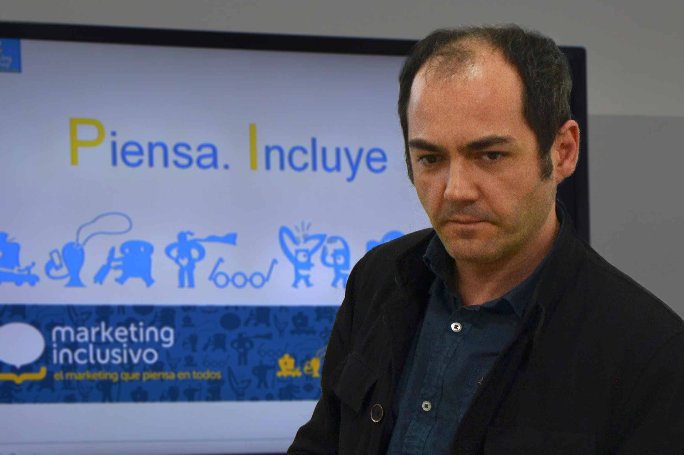 Luis posa frente a una pantalla colgada de la pared en la que junto a personajes dibujados que reflejan la diversidad humana, se ve el logo de Marketing Inclusivo. Se leen las palabras: Piensa. Incluye.